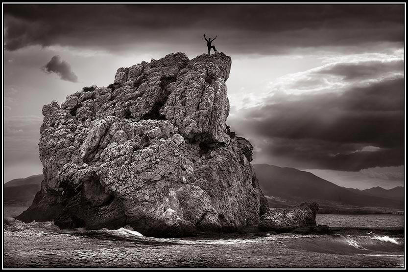 A Guy, arms upraised, atop a big dramatic rock.
